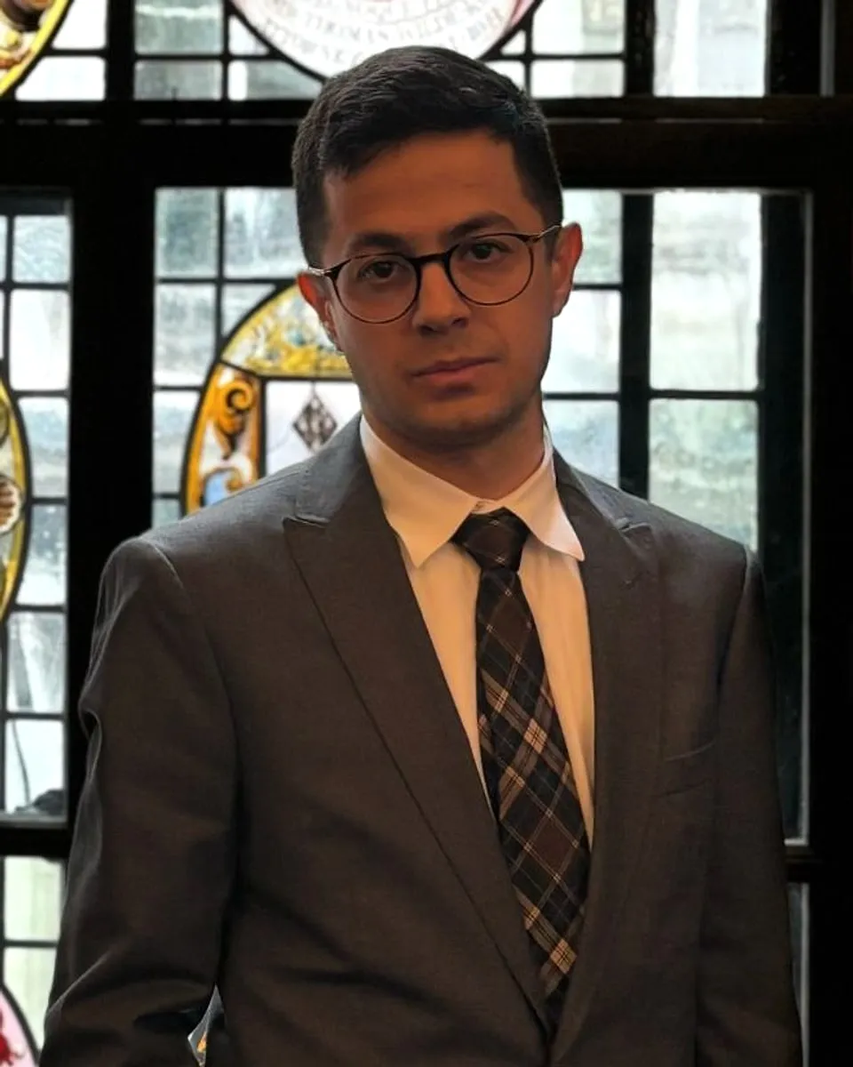 Sadik Bugra Isler, solicitor, wearing a grey suit and glasses, standing in front of a stained-glass window